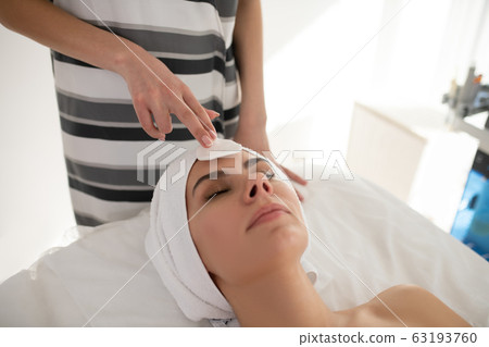 Cosmetologist in striped shirt cleaning clients face with cotton sponge 63193760