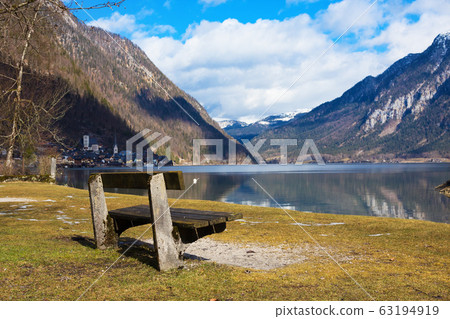Empty wooden bench on shore of beuatiful lake in austrian Alps. Winter sunny morning on lake Hallstatt. The most beautiful lake to explore in Austria. 63194919