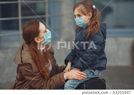 A European mother in a respirator with her daughter are standing near a building.The parent is teaching her child how to wear protective mask to save herself from virus 63203897
