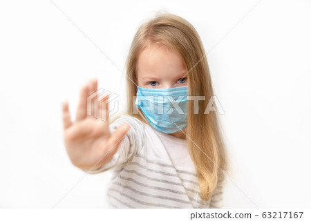 little girl putting up her hand, to say no, or stop, with a serious frowned expression in a medical mask on his face on a light background, close-up. 63217167