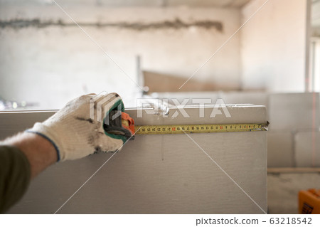 The construction worker measures with a tape measure Gypsum plate. Construction of internal walls in the apartment using a plaster concrete plate with groove ridge. tongue-and-groove gypsum blocks The construction worker measures with a tape measure Gypsum plate. Construction of internal walls in the apartment using a plaster concrete plate with groove ridge. tongue-and-groove gypsum blocks 63218542