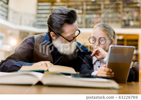 Smart pretty girl in eyeglasses and old man teacher having a lesson and discussion in vintage library. Girl holds digital tablet or ebook reader and man shows some information in book Smart pretty girl in eyeglasses and old man teacher having a lesson and discussion in vintage library. Girl holds digital tablet or ebook reader and man shows some information in book 63226769