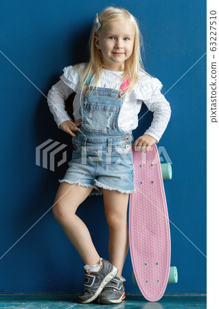Happy toddler girl with blond hair holding a skateboard while posing on blue wall background Happy toddler girl with blond hair holding a skateboard while posing on blue wall background 63227510