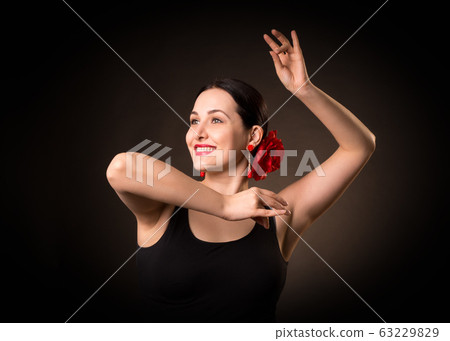 Female portrait of emotional flamenco dancer. Expressive smiling woman with hair up in a bun and red rose in black dress doing a pose. Isolated, low key. 63229829