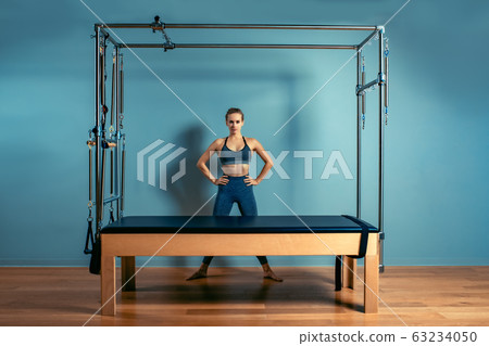 Young girl doing pilates exercises with a reformer bed. Beautiful slim fitness trainer on reformer gray background, low key, art light. Fitness concept 63234050