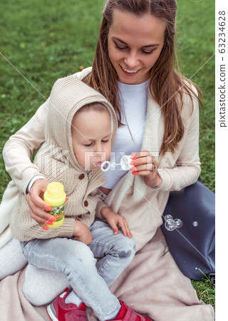 Happy woman mom, little boy plays inflates soap bubbles, casual warm clothes hood. Autumn day summer on grass. Weekend breaks, caring parenting childcare. Emotions Happiness joy fun smile. 63234628