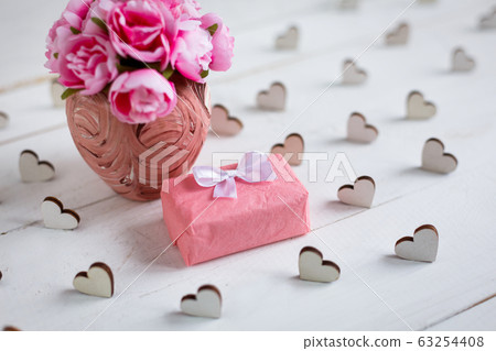 Small gift wrapped in paper with wooden hearts around and pink flowers in vase on white wooden background. Small gift wrapped in paper with wooden hearts around and pink flowers in vase on white wooden background. 63254408