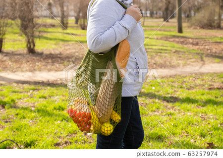 Female hand holds a green ecological cotton mesh bag with a bunch of organic fruits, vegetables and groceries: tomato, bananas, cheese and apples. Zero waste concept. Background, copy space, close up. Female hand holds a green ecological cotton mesh bag with a bunch of organic fruits, vegetables and groceries: tomato, bananas, cheese and apples. Zero waste concept. Background, copy space, close up. 63257974
