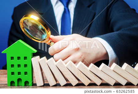 A businessman is studying the consequences of a domino falling on a house. Stress resistance of business and structures. Reliability and quality. Analysis. Real estate insurance and risk management. A businessman is studying the consequences of a domino falling on a house. Stress resistance of business and structures. Reliability and quality. Analysis. Real estate insurance and risk management. 63262585