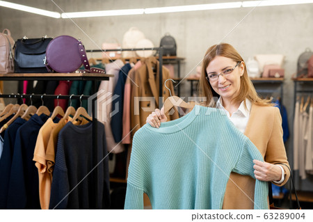 Happy elegant female in coat and eyeglasses showing you blue cotton sweater 63289004