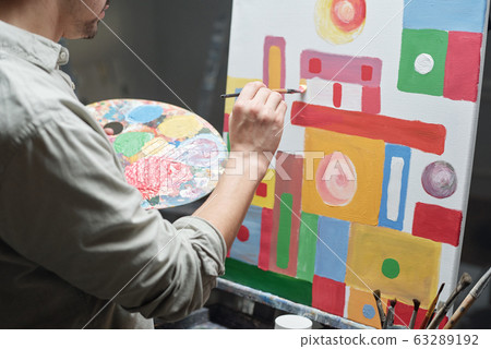 Young male artist with color palette and paintbrush working over painting 63289192