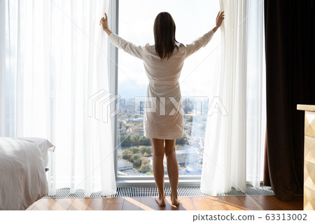 Young slim brunette woman opening curtains in morning. Young slim brunette woman opening curtains in morning. 63313002
