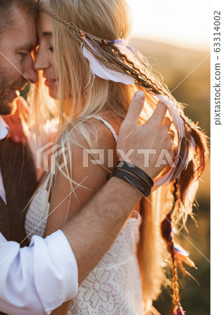 Close up portrait of beautiful boho couple embracing, at the sunset in summer field. Girl in white summer dress and feathers in hair, man in stylish casual suit. Summer vacation concept, boho couple Close up portrait of beautiful boho couple embracing, at the sunset in summer field. Girl in white summer dress and feathers in hair, man in stylish casual suit. Summer vacation concept, boho couple 63314002