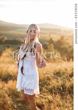 Happy attractive blond boho woman in white dress and feather accessories in hair in summer field outdoors, sunset. Boho fashion, hippie style Happy attractive blond boho woman in white dress and feather accessories in hair in summer field outdoors, sunset. Boho fashion, hippie style 63314038