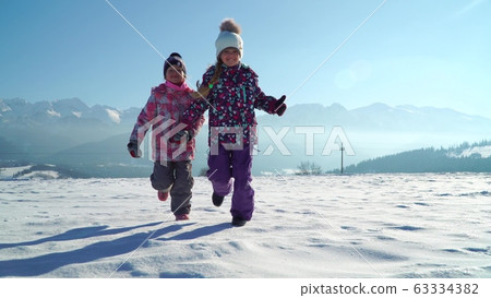 Charming kids in outwear running on snowy terrain with beautiful mountains on background in sunshine Charming kids in outwear running on snowy terrain with beautiful mountains on background in sunshine 63334382