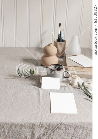 Artistic wokspace, still life. Paint brushes, pencils, olive branches, vase, cup of coffee and blank paper card mockups on linen tablecloth. Art supplies scene. Creative table background, vertical. Artistic wokspace, still life. Paint brushes, pencils, olive branches, vase, cup of coffee and blank paper card mockups on linen tablecloth. Art supplies scene. Creative table background, vertical. 63339827