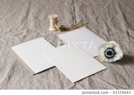 Wedding stationery mock-up scene. Blank greeting cards on linen tablecloth background with white anemone flower and silk ribbon. Feminine still life table composition. High angle view, no people. Wedding stationery mock-up scene. Blank greeting cards on linen tablecloth background with white anemone flower and silk ribbon. Feminine still life table composition. High angle view, no people. 63339843