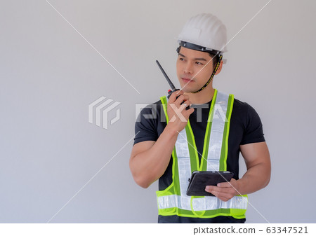 Strong foreman wearing a white helmet, holding a radio communication and a tablet on the construction site. Strong foreman wearing a white helmet, holding a radio communication and a tablet on the construction site. 63347521