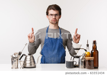 Barista, cafe worker and bartender concept. Portrait of upset grumpy and displeased young male employee feeling unsatisfied and jealous pointing up while standing working bar counter, make coffee Barista, cafe worker and bartender concept. Portrait of upset grumpy and displeased young male employee feeling unsatisfied and jealous pointing up while standing working bar counter, make coffee 63351434