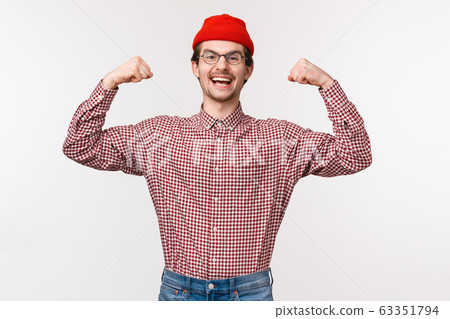 Waist-up portrait of encouraged and energized young confident slim guy in red beanie and glasses flex biceps, showing his muscles being strong and powerful, standing white background 63351794