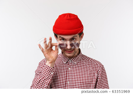 Close-up portrait of suspicious funny young guy in red beanie and checked shirt, take-off sunglasses and look from under forehead as gossiping, hinting or telling secret, spying on someone Close-up portrait of suspicious funny young guy in red beanie and checked shirt, take-off sunglasses and look from under forehead as gossiping, hinting or telling secret, spying on someone 63351969