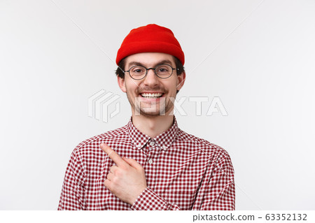 Close-up portrait cheerful handsome caucasian hipster guy in red beanie and glasses, pointing finger upper left corner and smiling pleased, showing advertisement, recommend product, special offer 63352132