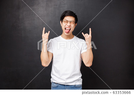 Happy excited and outgoing asian guy in glasses and t-shirt, having fun show tongue close eyes relaxed and make rock-n-roll heavy metal gesture as listening to awesome song, black background Happy excited and outgoing asian guy in glasses and t-shirt, having fun show tongue close eyes relaxed and make rock-n-roll heavy metal gesture as listening to awesome song, black background 63353666