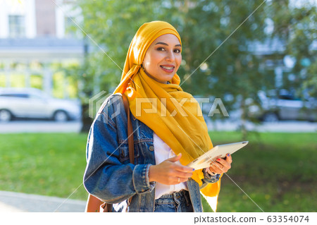 Arab woman student. Beautiful muslim female student wearing bright yellow hijab holding tablet. 63354074