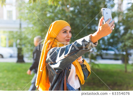 Muslim girl in hijab makes a selfie on the phone standing on the street of the city Muslim girl in hijab makes a selfie on the phone standing on the street of the city 63354076
