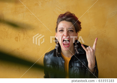 Let's rock concept. Portrait of excited beautiful woman with dark pink hair in casual yellow dress standing, looking at camera, showing rock and roll gesture. Let's rock concept. Portrait of excited beautiful woman with dark pink hair in casual yellow dress standing, looking at camera, showing rock and roll gesture. 63355183