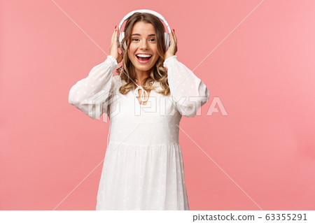 Portrait of excited, happy good-looking girl in white tender dress, wearing headphones and smiling amazed as looking at camera, fascinated with good sound quality, pink background Portrait of excited, happy good-looking girl in white tender dress, wearing headphones and smiling amazed as looking at camera, fascinated with good sound quality, pink background 63355291