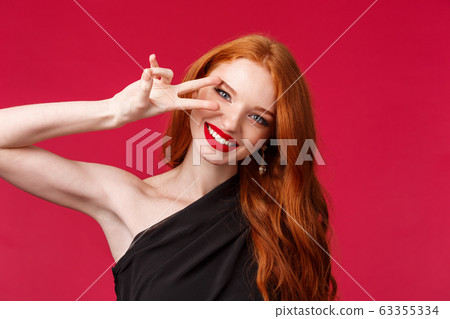 Elegance, beauty and fashion concept. Close-up portrait of gorgeous seductive redhead woman, caucasian female model in black dress, makeup and red lipstick, show peace sign near eye, smiling 63355334