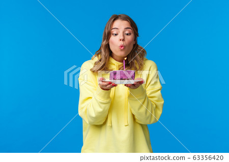 Celebration, party and lifestyle concept. Silly cute european blond woman in yellow hoodie, making wish holding fruit cake blowing out b-day candle, standing blue background at birthday party 63356420