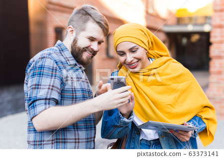 Handsome man shows a photo on smartphone young arab woman. Beautiful muslim female student wearing bright yellow hijab communicates with man outdoors. 63373141
