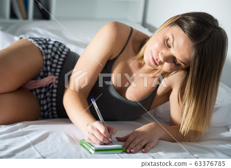 Young smiling girl lounging in bed and writing to notebook 63376785