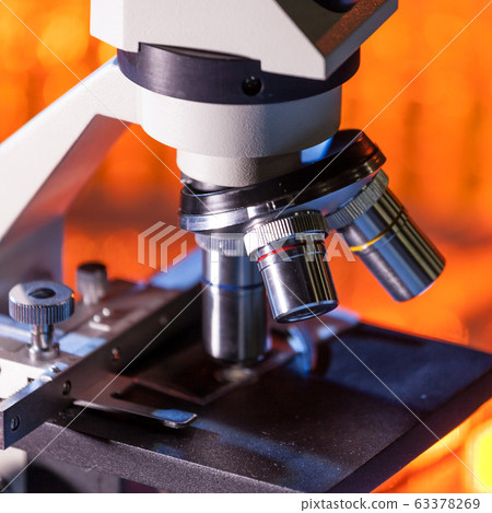 Close up of microscope lenses focused on a specimen in warm orange light light. Close up of microscope lenses focused on a specimen in warm orange light light. 63378269