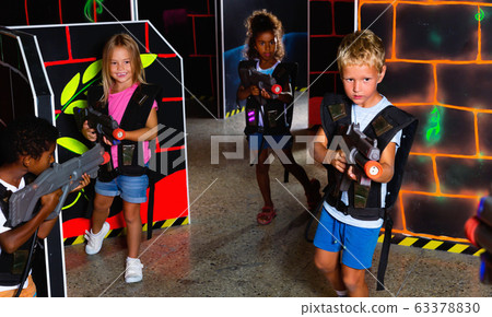 Multiracial team of happy tween kids aiming laser guns at other players during lasertag game in dark room 63378830