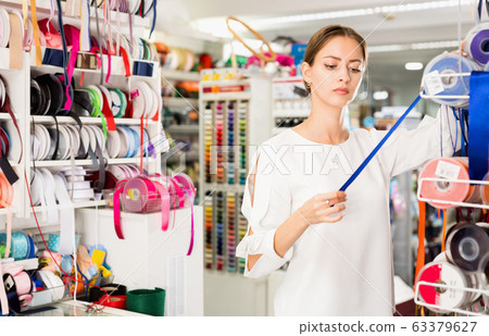 Interested young woman choosing colorful ribbons and braid for dressmaking in sewing supplies shop 63379627