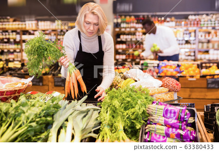 Experienced middle aged saleswoman working in organic food store, arranging fresh vegetables on counter 63380433