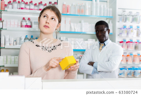 Pensive young woman choosing medicines in modern pharmacy Pensive young woman choosing medicines in modern pharmacy 63380724