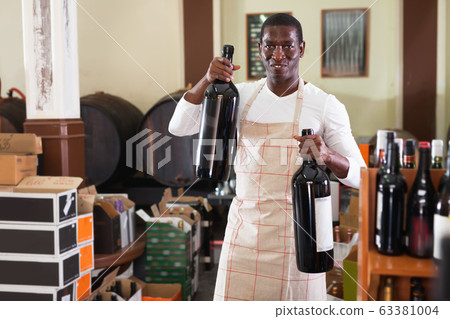 Smiling salesman in apron proposing wine in bottles in winery shop Smiling salesman in apron proposing wine in bottles in winery shop 63381004