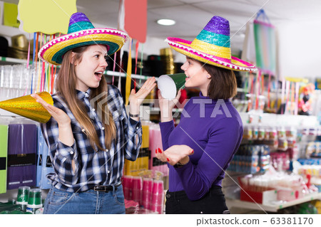 Smiling attractive girls trying on party hats in accessories shop Smiling attractive girls trying on party hats in accessories shop 63381170