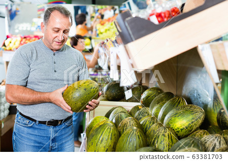Smiling male is choosing green melon in the supermarket. Smiling male is choosing green melon in the supermarket. 63381703
