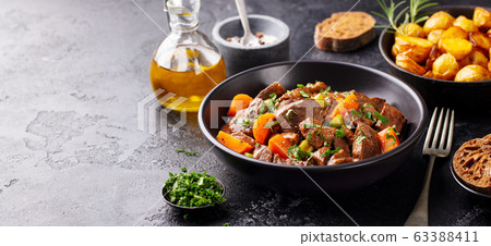 Beef meat and vegetables stew in black bowl with roasted baby potatoes. Dark background. Copy space. 63388411