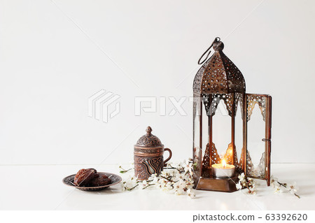 Ramadan Kareem greeting card, invitation. Bronze plate with dates fruit, white flowers, prunus tree blossoms, cup of tea and glowing Moroccan lantern on table background. Iftar dinner. Eid ul Fitr. 63392620