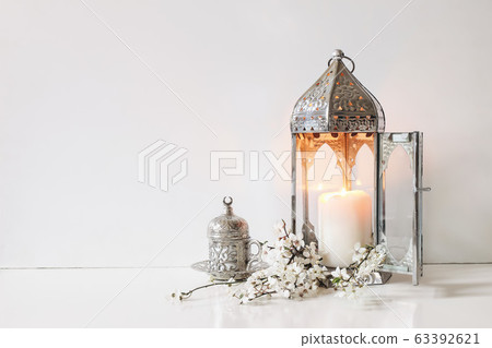 Silver decorative cup of tea, white flowers, prunus tree blossoms and glowing Moroccan lantern on table background. Iftar dinner. Ramadan Kareem greeting card, invitation. Eid ul Fitr muslim holiday. Silver decorative cup of tea, white flowers, prunus tree blossoms and glowing Moroccan lantern on table background. Iftar dinner. Ramadan Kareem greeting card, invitation. Eid ul Fitr muslim holiday. 63392621