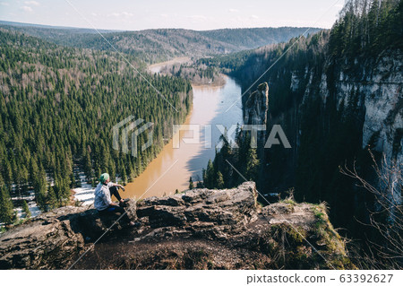 Man traveller sitting near edge of cliff looking at beautiful spring landscape Man traveller sitting near edge of cliff looking at beautiful spring landscape 63392627