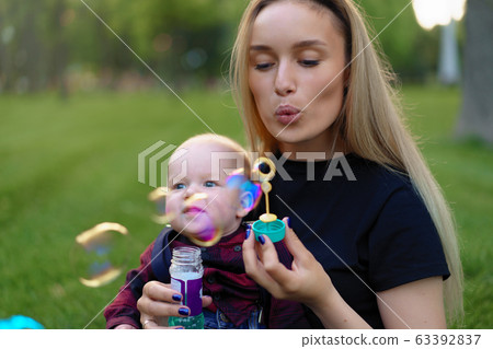 Young Caucasian mother inflates soap bubbles with her little son in a park on a sunny summer day. 63392837