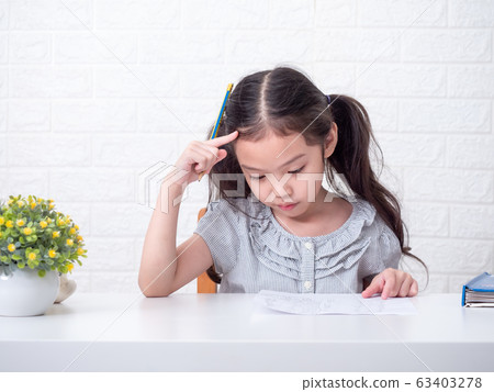 Asian little cute girl 6 years old seriously thinking about math homework over white brick wall background and white table. Learning and education Asian little cute girl 6 years old seriously thinking about math homework over white brick wall background and white table. Learning and education 63403278