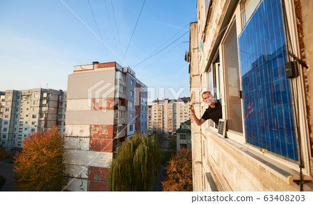 Man waves on the balcony of multi-storey building equipped with solar panel, urban background Man waves on the balcony of multi-storey building equipped with solar panel, urban background 63408203